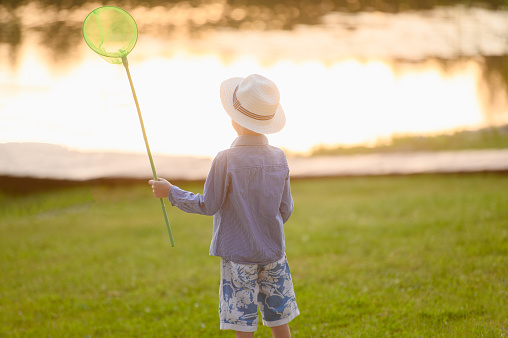 A boy in a hat at sunset plays with scoop-net outdoors on a summer day. Summer outdoor activities for children, exploring nature, traveling, family vacation.