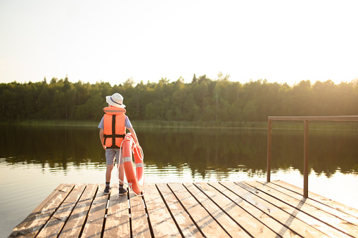 A boy in a life jacket, a hat and a life buoy stands on a pier near the water in the rays of the setting sun. Water safety concept.Copy space