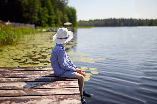 A boy in a hat sits on a wooden pier by the lake on a summer day. The boy dreams looking into the distance. The concept of childhood, solitude with nature, dreams. Copy space.