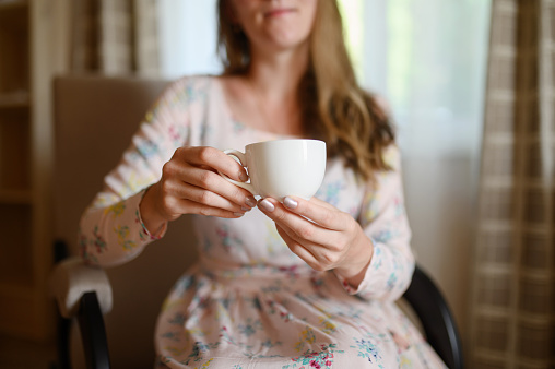 Young woman drinking tea or coffee holding a white cup with both hands while relaxing in a rocking chair. White mug mockup. The concept of rest on the weekend, on vacation.