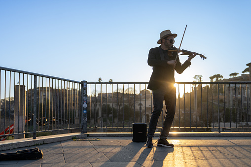 An elegant street musician plays the violin for people passing by him in Rome, a few meters from the Colosseum. The violinist has a very elegant style dressed in a suit and hat and the photograph taken at sunset shows him against the light.