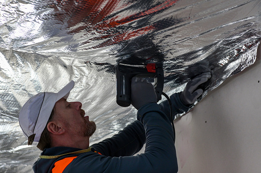 a worker in a vest and cap is fixing the vapor barrier on the ceiling.