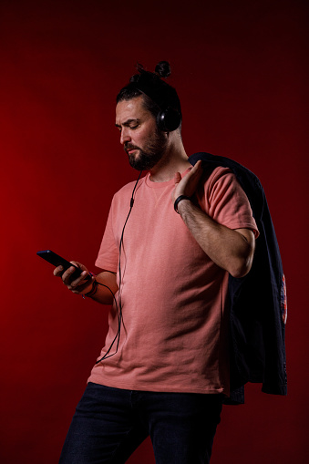 Portrait of mid adult man standing against bright red background, holding his blazer on the shoulder and scrolling through his playlist on smart phone while listening to music via headphones.