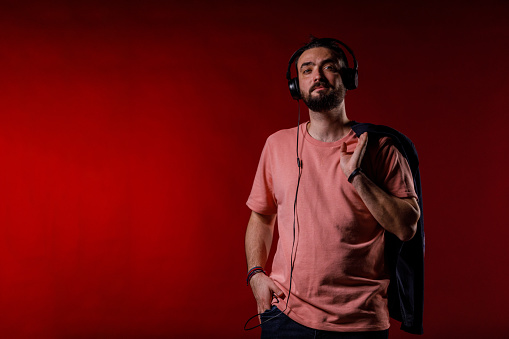 Copy space shot of cool mid adult hipster standing against bright red background, holding his jacket on the shoulder and listening to music via headphones. He is looking away, smiling and contemplating.