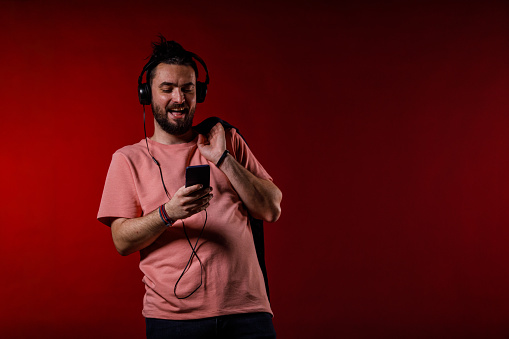 Man, smile and headphones for music on phone in studio by blue background. Asian person, listening and streaming a song, radio and audio or funny podcast on smartphone, technology and sound for joy