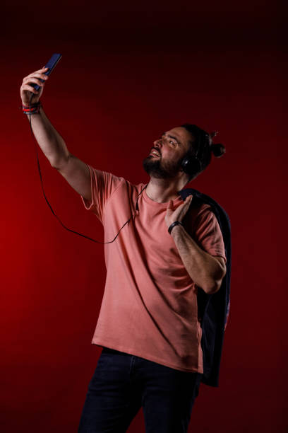 Portrait of mid adult man standing against bright red background, holding his blazer on the shoulder and scrolling through his playlist on smart phone while listening to music via headphones.