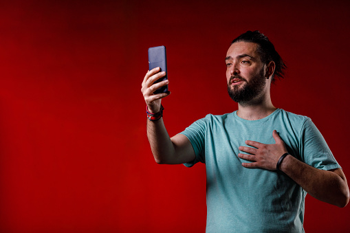 Portrait of charming hipster standing against bright red background, video chatting with a friend via smart phone.