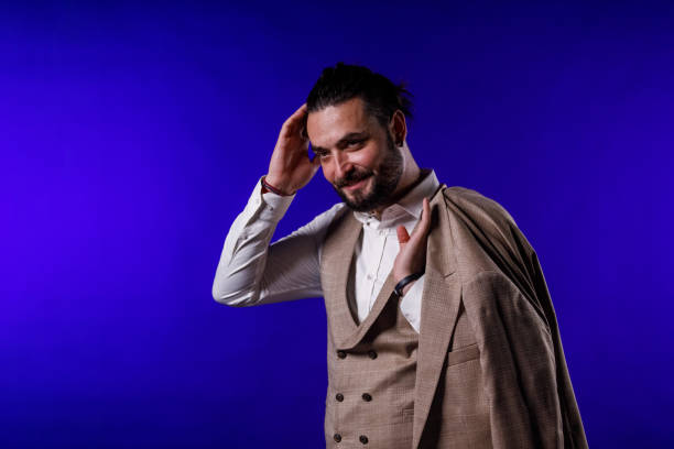 Portrait of charming mid adult man standing against bright blue background, holding his suit jacket on the shoulder, running his hand through his hair, smiling with confidence.