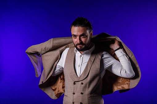 Portrait of charming mid adult man standing against bright blue background, putting on his suit jacket.