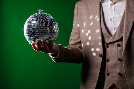 Midsection of unrecognizable, charming man in a suit, standing against dark green background and playing with a disco ball.