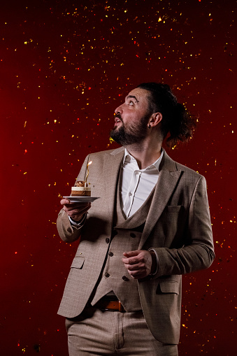 Portrait of charming mid adult man in a suit standing against bright red background, holding a small cake with a birthday candle and making a wish while confetti are exploding in the background.