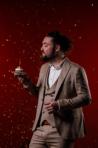 Portrait of charming mid adult man in a suit standing against bright red background, holding a small cake with a birthday candle and making a wish while confetti are exploding in the background.