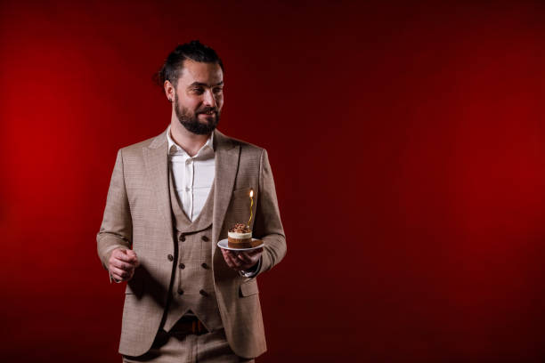 Portrait of charming mid adult man in a suit standing against bright red background, holding a small cake with a birthday candle and making a wish.