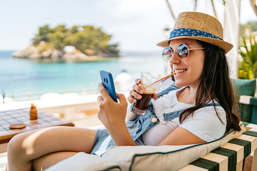Relaxed young woman using her phone and drinking iced coffee at a seaside café in Parga, Greece.