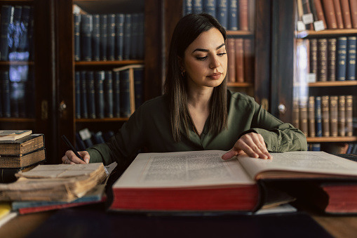 Young woman reading big old book and making a note while sitting in library in front of bookshelves. She wears green shirt and looks beautiful