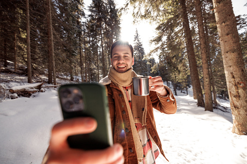 Young handsome man taking selfie while having coffee outdoors in nature on sunny day