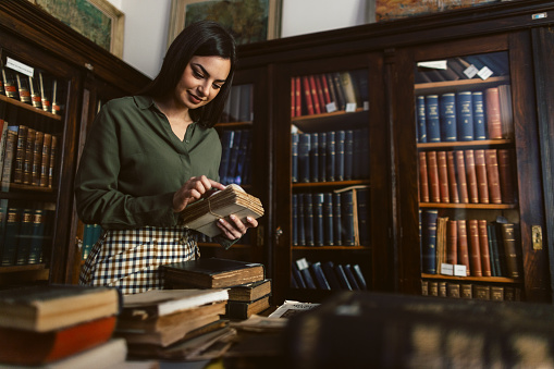 Young woman holding and carrying old scrolls while walking in library in front of bookshelves. She wears green shirt and she is unrecognizable