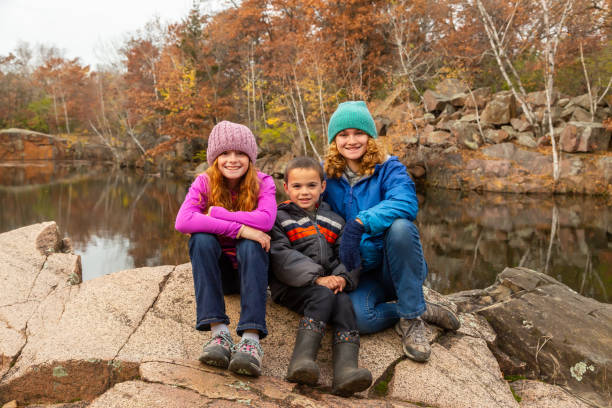 Two sisters and their younger brother smiling at the camera on an autumn day in Minnesota, USA. They are out for a hike and sitting at the edge of an old granite quarry pond.