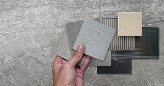 architect hold tiles swatch to compare with materials on board including corrugated glasses, grainy quartz, stone ceramic tiles, black marble placed on grey emperador marble table.