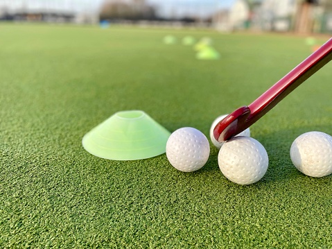 closeup of a red hockey stick with white balls and sport yellow cone next to it, on a green grass hockey field. Artificial grass for sports fields.