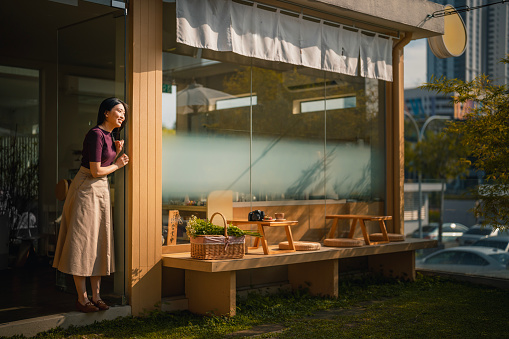 Asian Chinese woman restaurant owner standing in front of restaurant looking away smiling morning