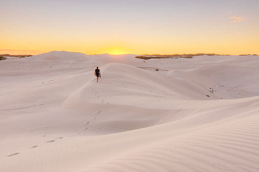 Young backpacker hiking through arid desert landscape scene with white sand dunes against a golden sky. Photographed in Western Australia.