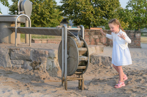 child operating a manual water pump at a sandy playground. childs face is obscured. playground is surrounded by greenery, and there are trees in the background under a clear sky.
