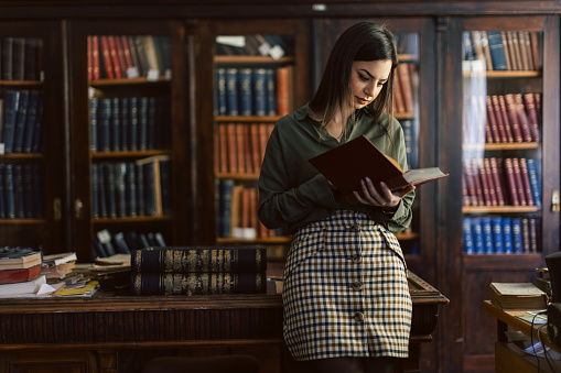 Dreamy happy young beautiful Caucasian woman looking in distance, distracted from studying on online courses, reading educational books or preparing for exams alone in modern college library.