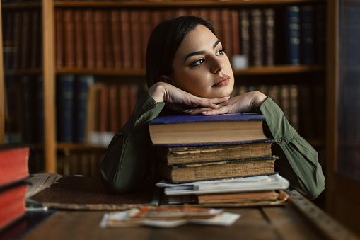 Young woman taking a break and looking away after packing and sorting old books while sitting in library in front of bookshelves. She wears green shirt and looks beautiful