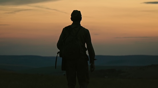 Silhouette of lonely soldier walking in field on red sunset background. A sad conceptual scene about the history of the soldiers of World War II and the Cold War. Cinematic video footage shot.