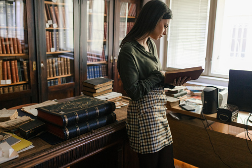 Young woman holding old book while standing in library in front of bookshelves. She wears green shirt and looks beautiful