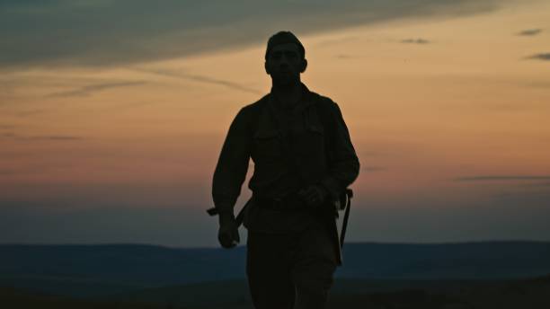 A lone soldier with a backpack from World War II returns home from the war walking through a summer field against a red sunrise. Cinematic video footage shot.