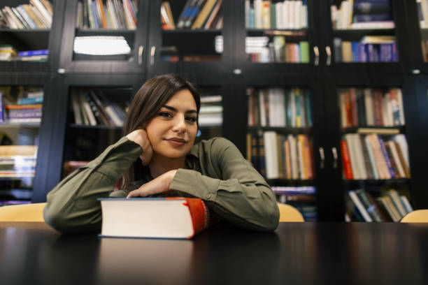 Young woman taking a break and looking away after reading big old book while sitting in library in front of bookshelves. She wears green shirt and looks beautiful