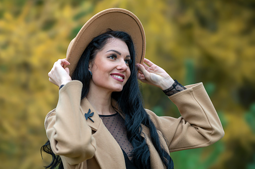 Beautiful brunette woman in beige coat and hat posing in autumn park