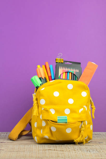 istock Yellow backpack with white polka dots with different colorful stationery on table. Purple background. Back to school. 2003245516
