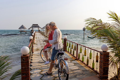 Mature couple ride tandem bicycle along pier towards straw umbrellas over the sea