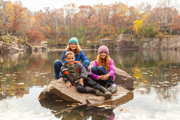 Two sisters and their younger brother sitting on a large rock in an old granite quarry pond. Taken on an autumn day at Quarry Park in Minnesota, USA.