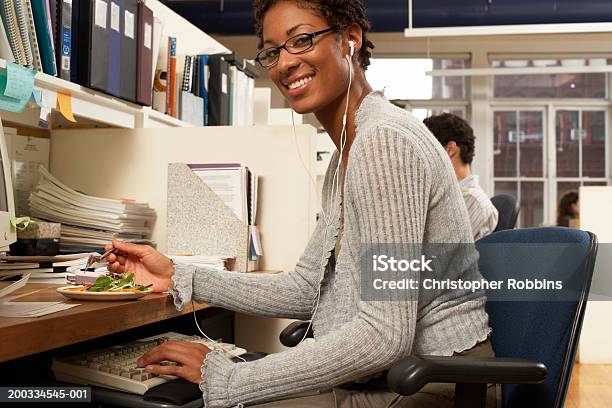 Mujer Comiendo En Escritorio De Oficina Sonriendo Retrato Foto de stock y más banco de imágenes de Auriculares - Equipo de música