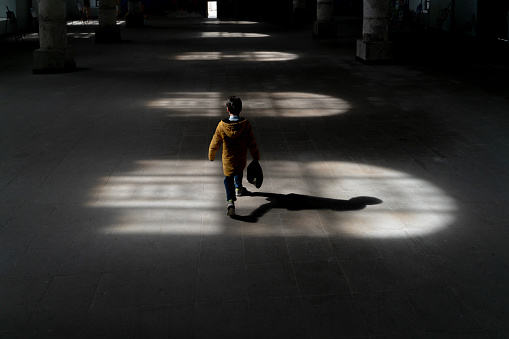 Lights hitting the floor from the window inside the building and a boy walking in the light. The child walks with his head down. A photo with shadows. Taken indoors in daylight with a full frame camera.