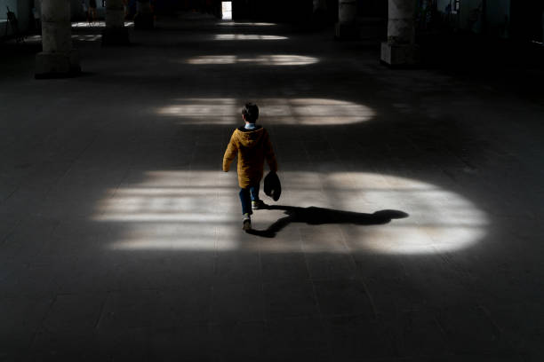 Lights hitting the floor from the window inside the building and a boy walking in the light Lights hitting the floor from the window inside the building and a boy walking in the light. The child walks with his head down. A photo with shadows. Taken indoors in daylight with a full frame camera. silhouette of bad boy stock pictures, royalty-free photos & images