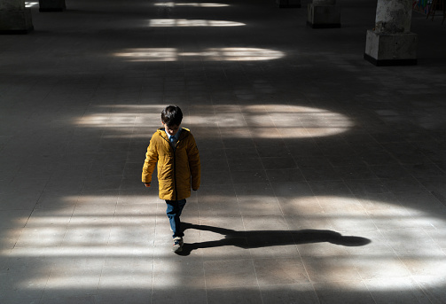 Lights hitting the floor from the window inside the building and a boy walking in the light. The child walks with his head down. A photo with shadows. Taken indoors in daylight with a full frame camera.