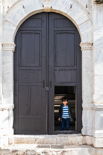 Boy looking out from the large entrance door of the church. The boy is standing on the open part of the large wooden door of the church. Taken in daylight with a full frame camera.