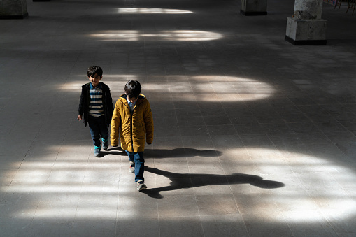Lights hitting the ground from the window inside the building and a child walking in the light, his brother right behind him, smiling and looking at the camera. The child walks with his head down. A photo with shadows. Taken indoors in daylight with a full frame camera.