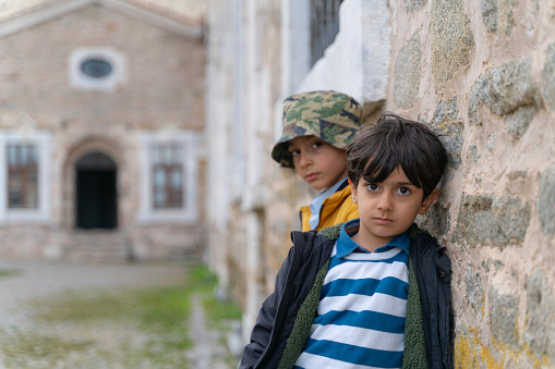 portrait photo of brothers leaning against the stone wall of the church. The little boy is photographed clearly in the foreground, with his older brother blurred behind him. Behind them is the entrance of the church. Taken in daylight with a full frame camera.