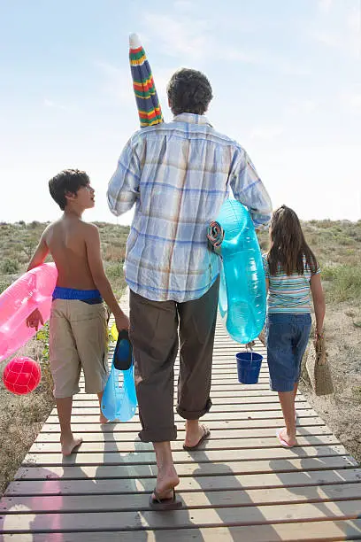 Photo of Father and children (7-9) on path carrying beach toys, rear view