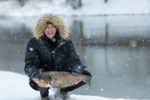 A fisherman on winter fishing caught a large silver carp and holds it in his hands. He rejoices at the good catch, despite the cold and snowfall.