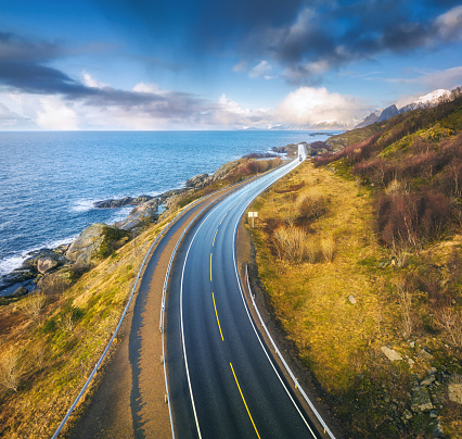 Aerial view of road, rocky sea coast with waves and stones at sunset in Lofoten Islands, Norway. Landscape with beautiful road, transparent blue water, rocks. Top view from drone of highway in summer