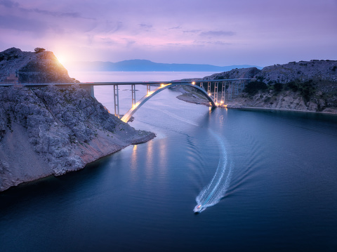 Aerial view of bridge over the sea at sunset. Modern Krk bridge with illumination, island, mountains, blue sea, boat and purple sky at night. Top drone view of road, city lights, rocks. Architecture