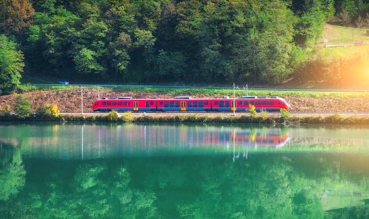 Beautiful red modern train and river in alpine mountains at sunrise in summer. Passenger train, reflection in water, railroad, lake, green forest in spring. Railway station in Slovenia