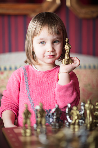 A little girl is playing chess indoor. She is thinking about her next move, holding the knave.